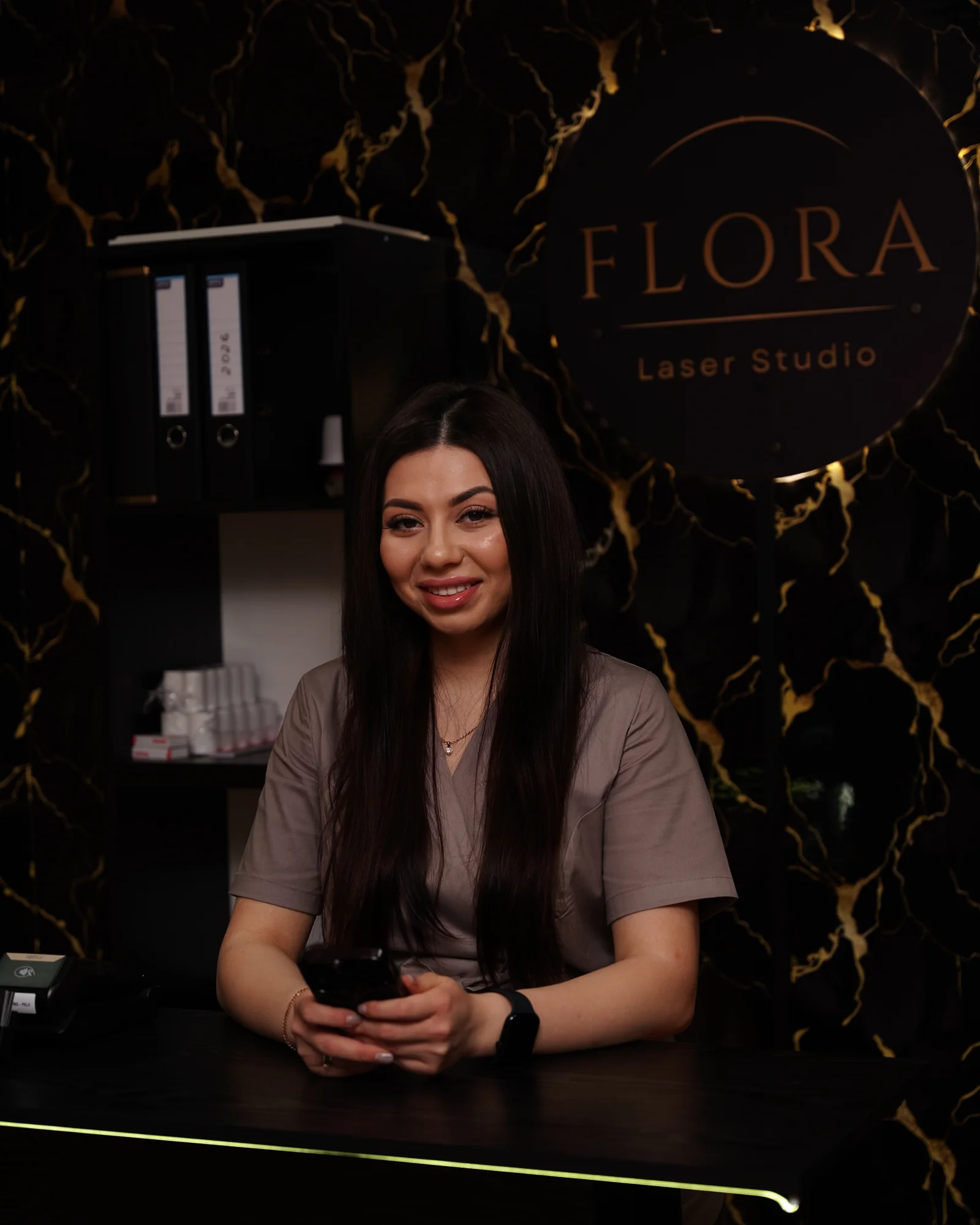 Flora przy ladzie recepcji, za nią podświetlony szyld FLORA Laser Studio na czarnym marmurze